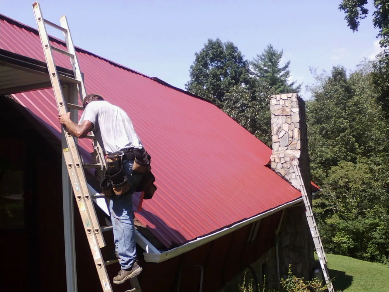 Roofer on a ladder beside a red metal roof and stone chimney — Soffit & Fascia Repair by Roofing Brothers in Flower Mound